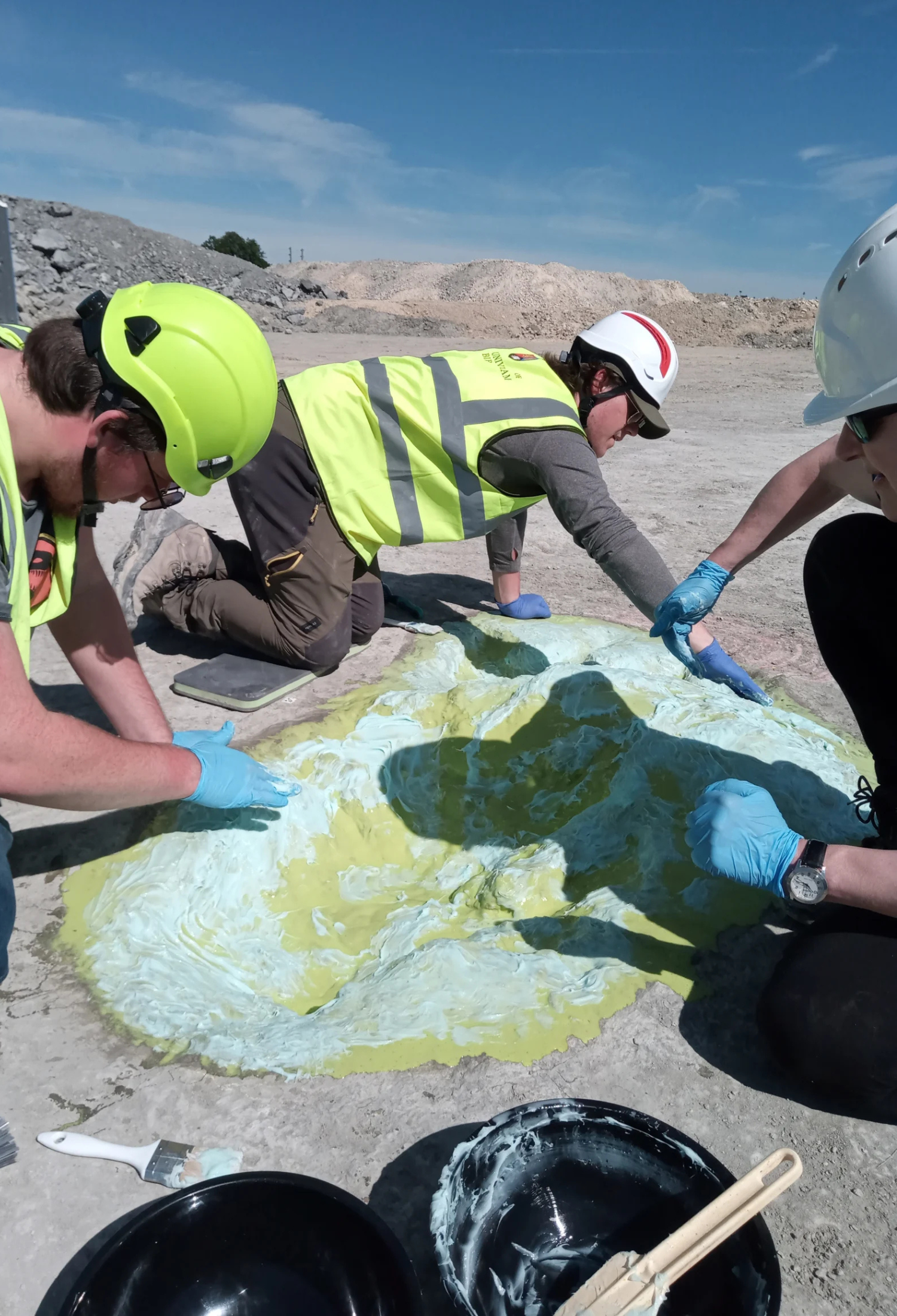 Three people wearing hard hats, fluorescent jackets and latex gloves apply a viscous sticky substance to one of the dinosaur footprints.