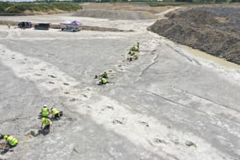 Aerial photograph showing volunteers in hard hats and fluorescent jackets digging out a series of dinosaur footprints in a quarry.