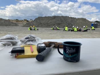 Foreground: a table with excavation equipment, sample bags and a mug saying 'Hit the Road.' Background: Volunteers digging out the footprints on the quarry floor.