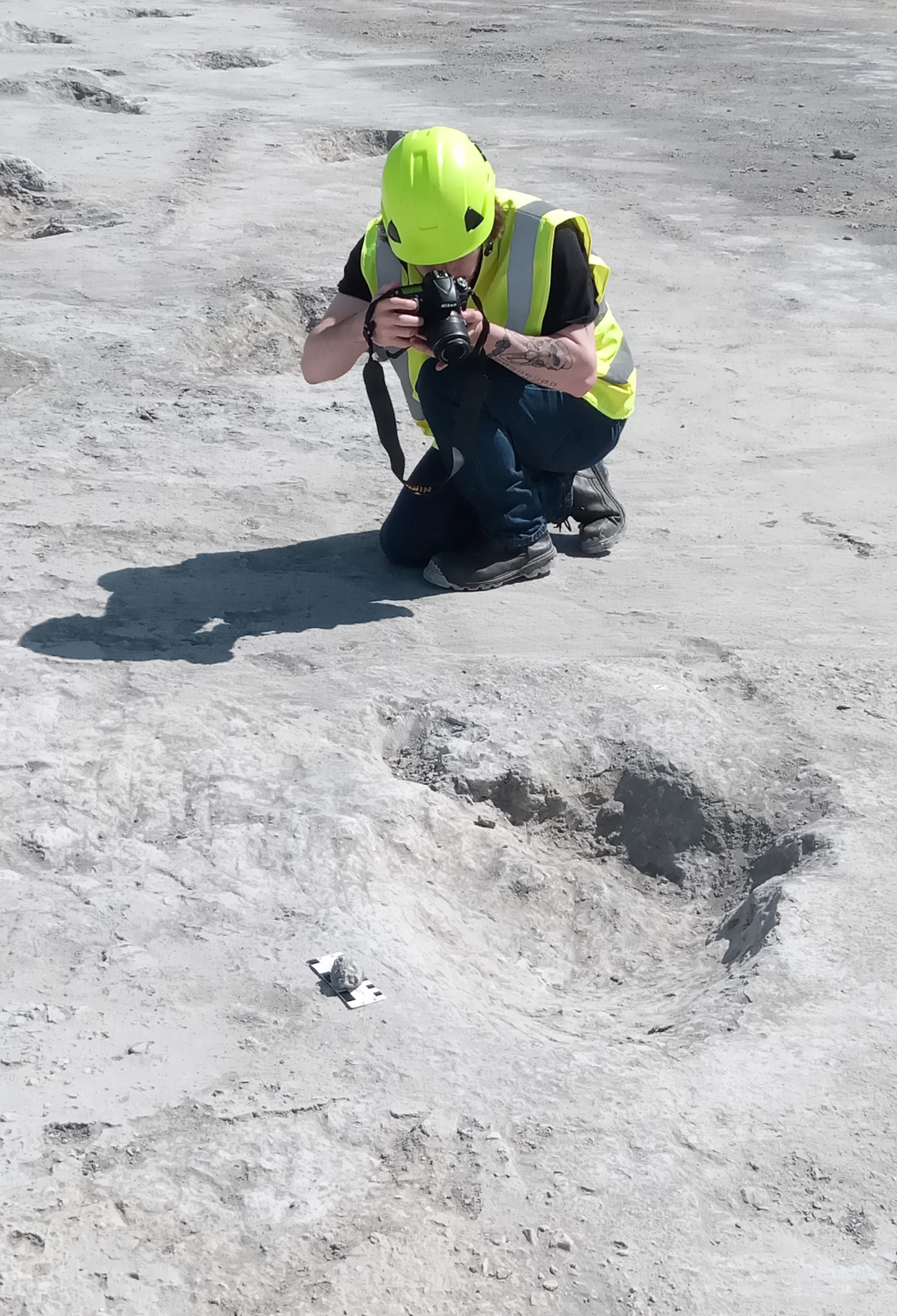 A young man wearing a hard hat and fluorescent jacket crouches next to one of the dinosaur footprints. He is taking a photograph of it using a DSLR camera.