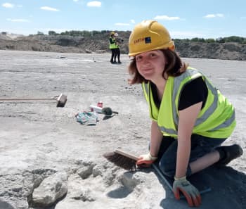 A young white lade with shoulder length brown hair wearing a hard hat and fluorescent jacket. She kneels at the side of a dinosaur footprint in the ground and holds a brush.