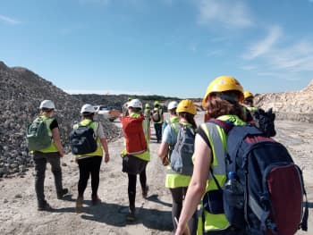 A group of people viewed from behind at a quarry. They all wear hard hats and fluorescent jackets, and wear rucksacks. It is a hot sunny day.