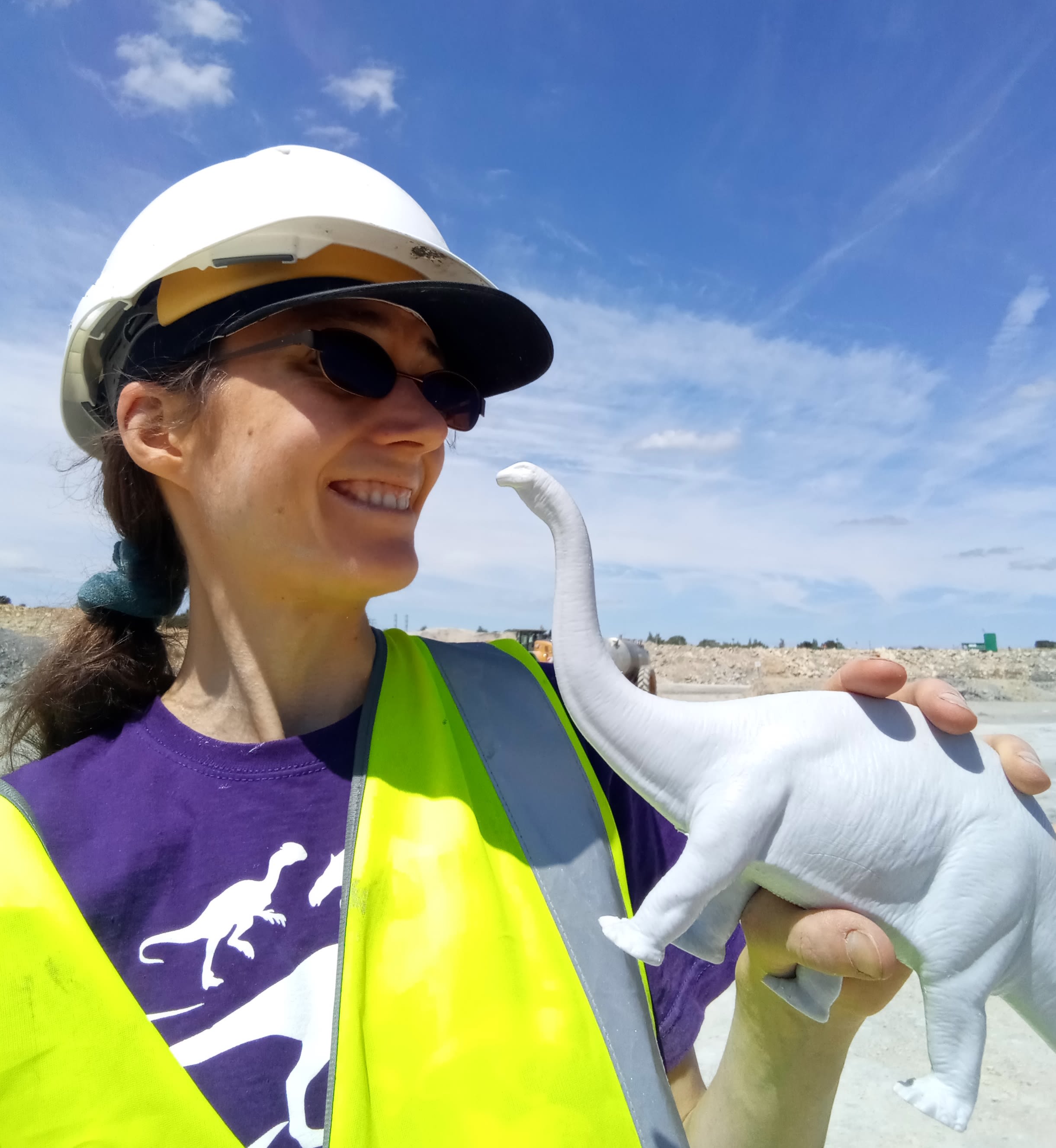 A young white woman with long brown hair tied back in a pony tail stands in a quarry. She wears a white hard hat and a flourescent jacket. She holds a plastic model of a diplodocus