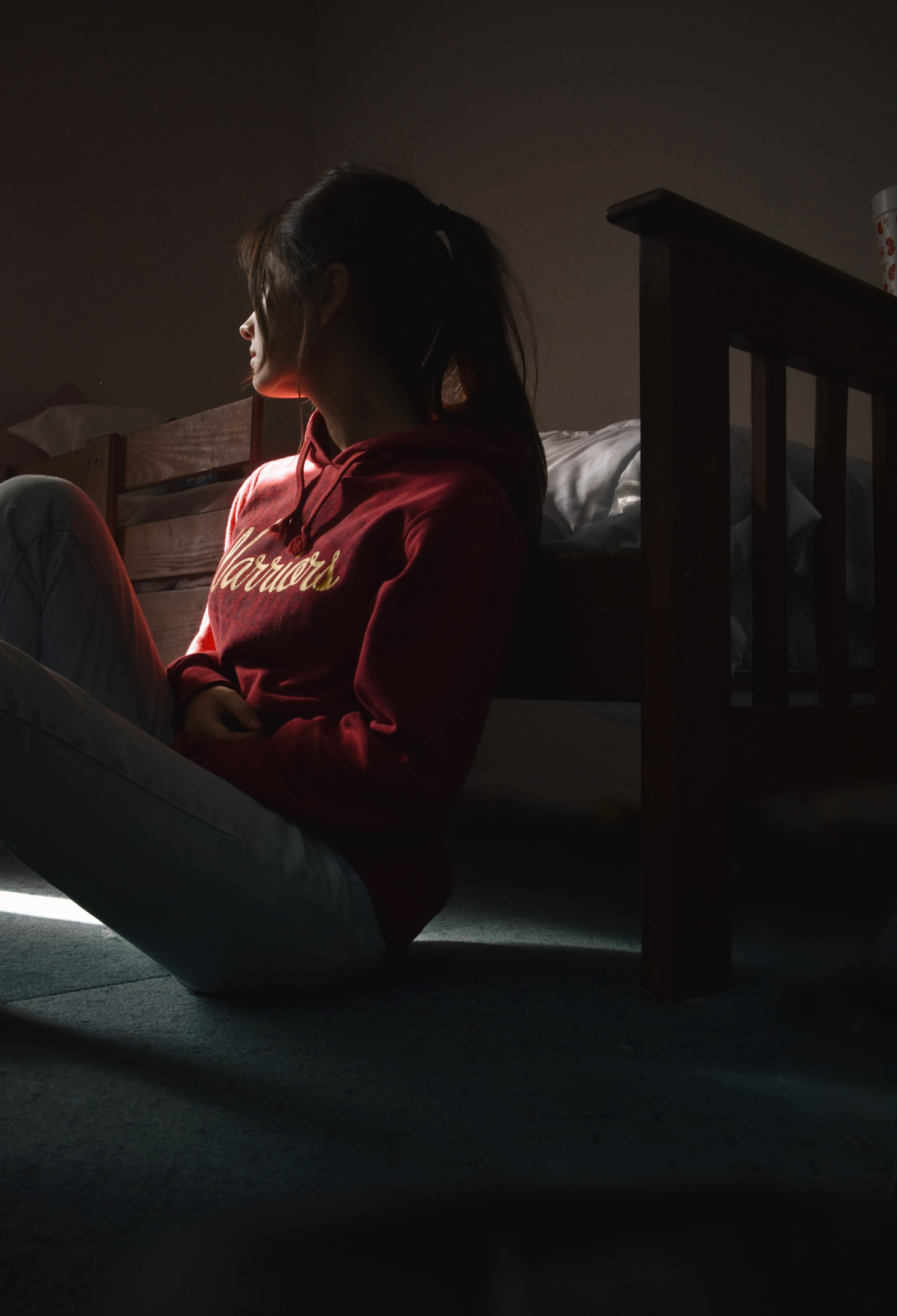 A young person leans against their bed and looks into the distance