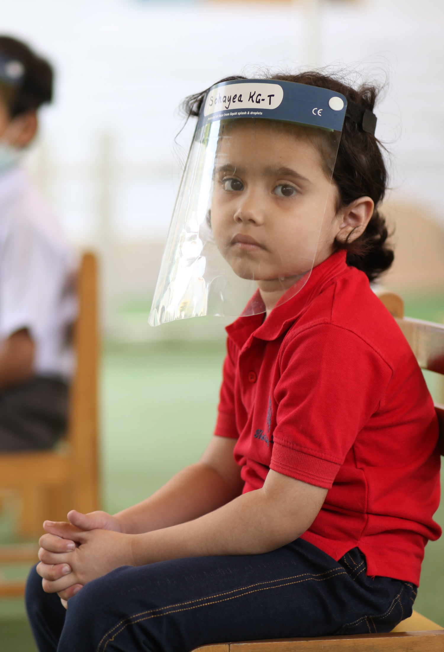 A young child sits socially distanced on a chair wearing a protective visor