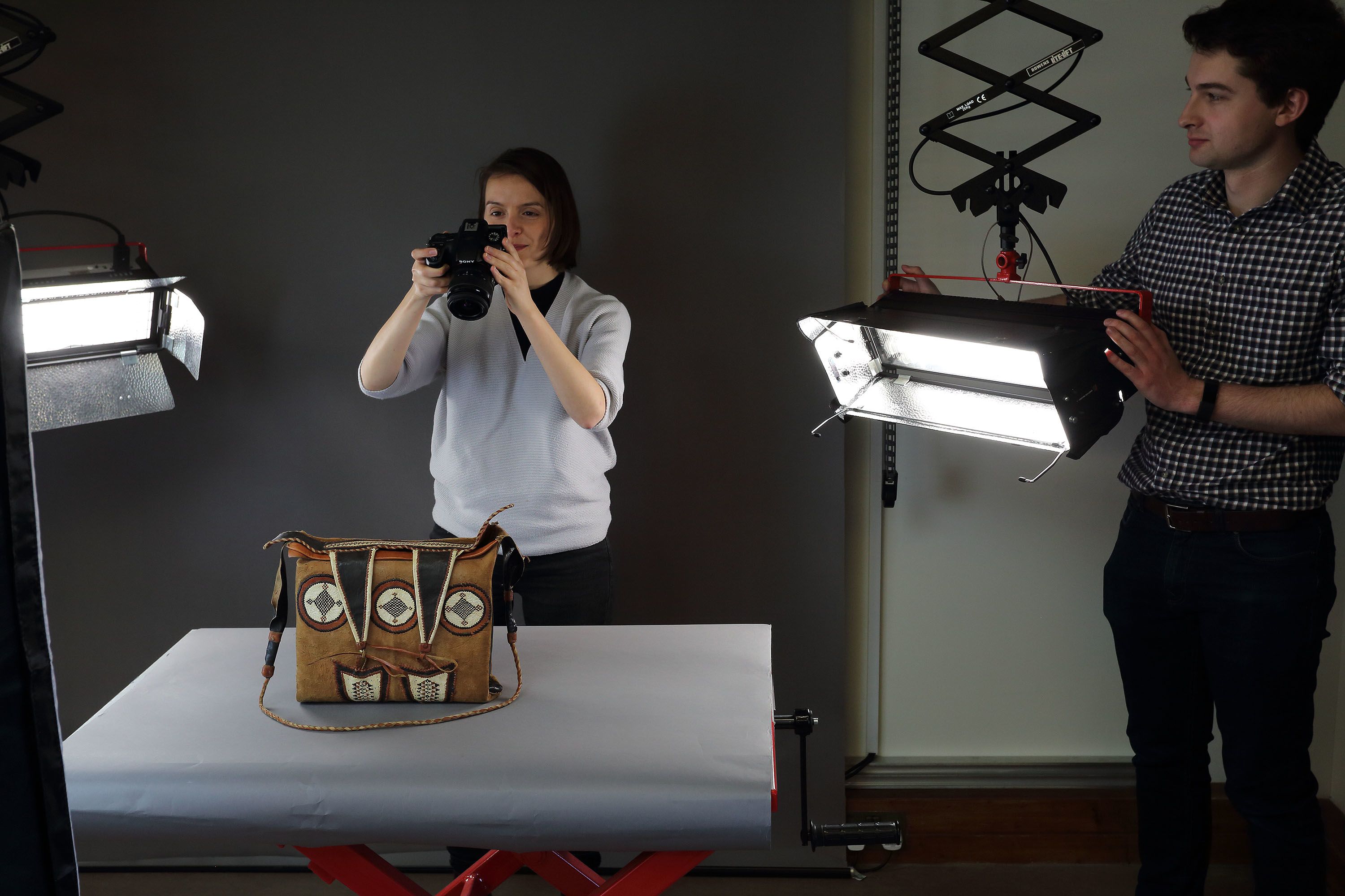 A woman photographing a satchel while a man holds a light