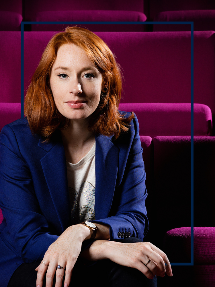 A portrait photo of Professor Hannah Fry, sat down in a row of chairs in a theatre