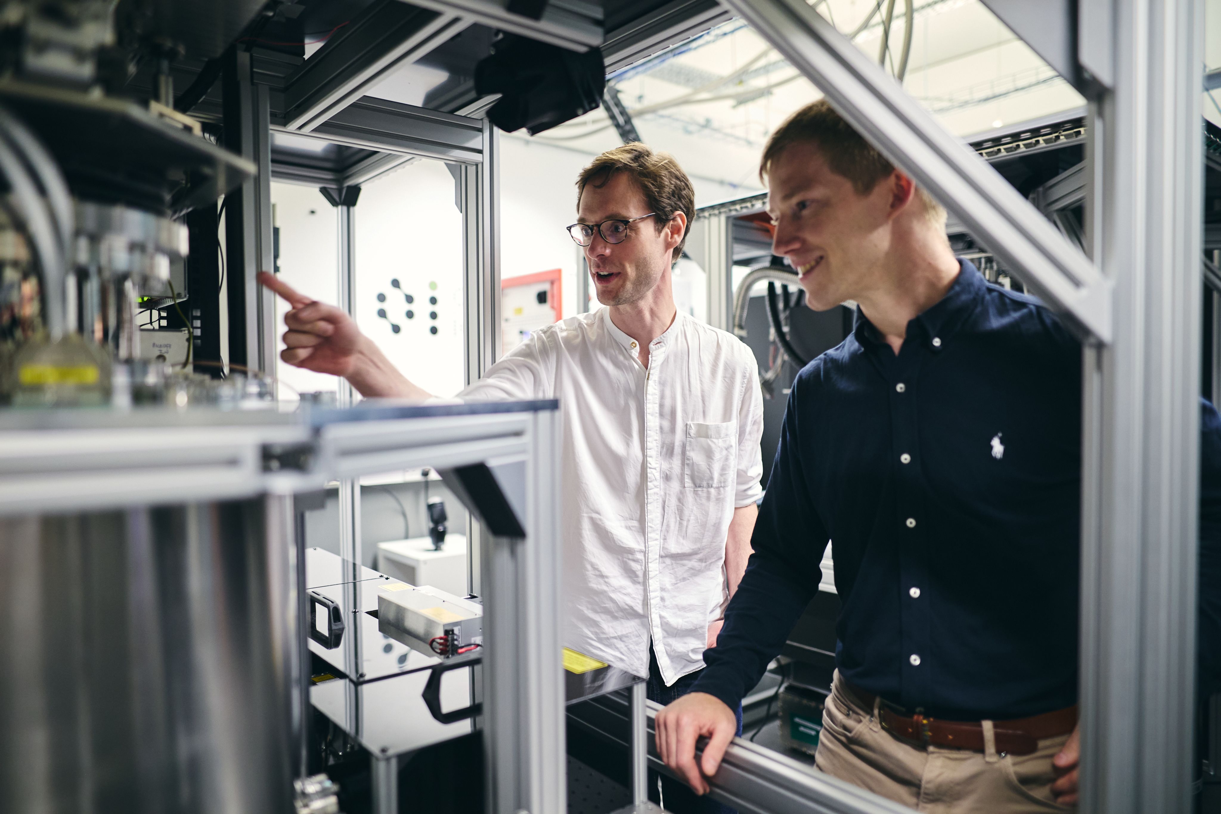 Two white men stand together with one pointing at the components of a quantum computer seen as an assembly of wires and cylinders.