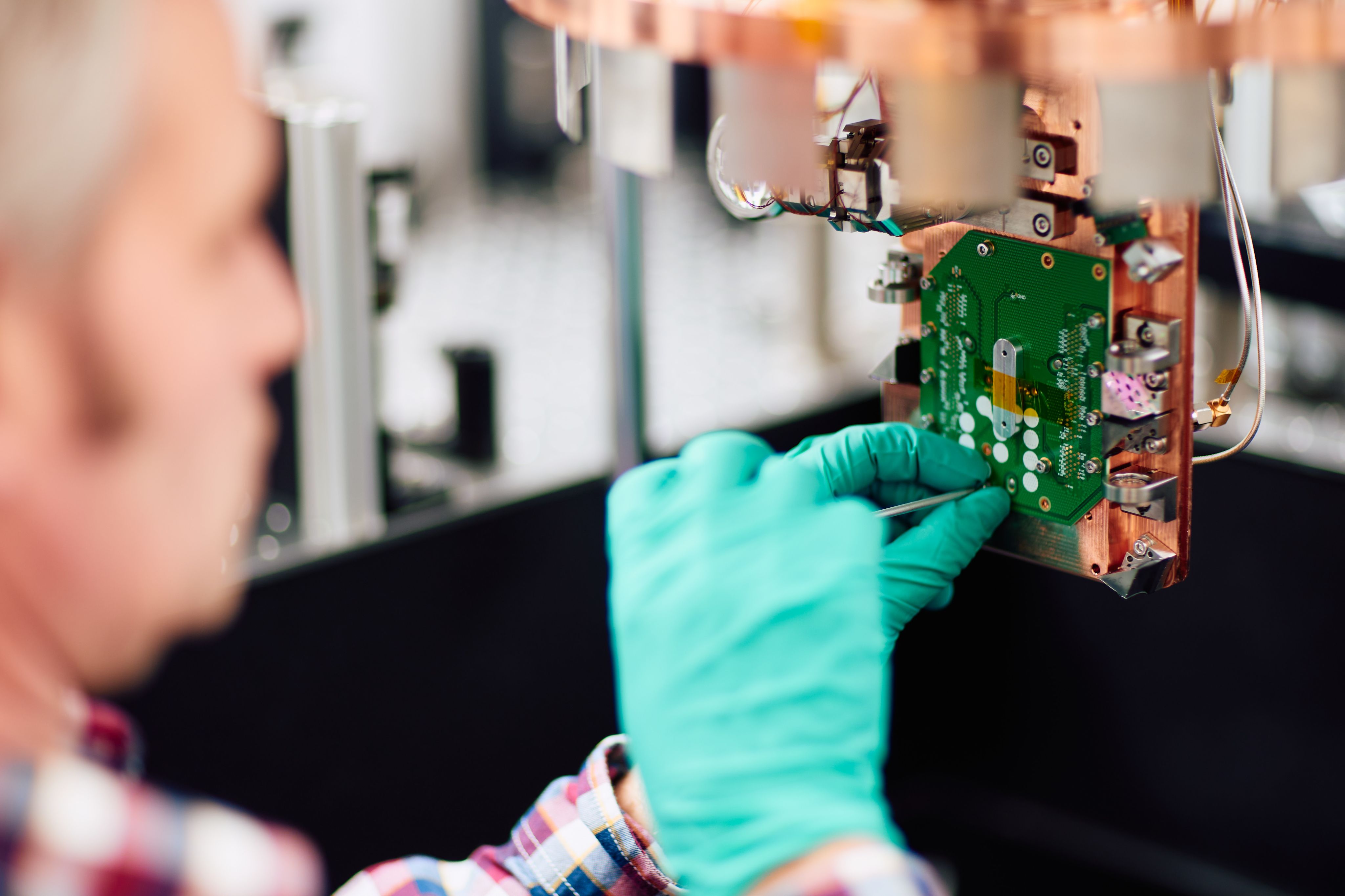 Close up of a man wearing green latex gloves who is working on a component of a quantum computer with a screwdriver.