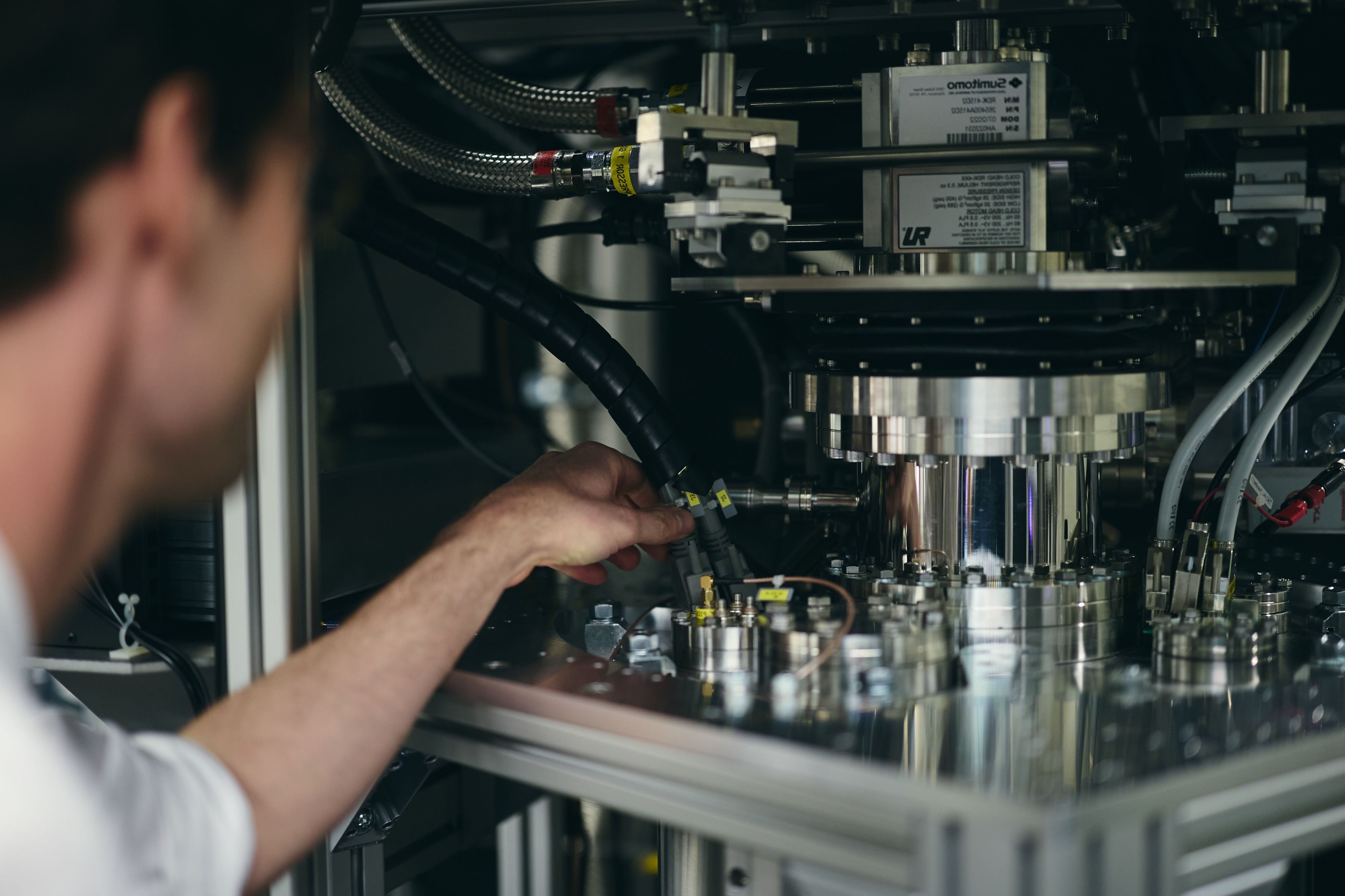 A white man faces away from the viewer and reaches towards the components of a quantum computer seen as an assembly of buttons, wires and cylinders.