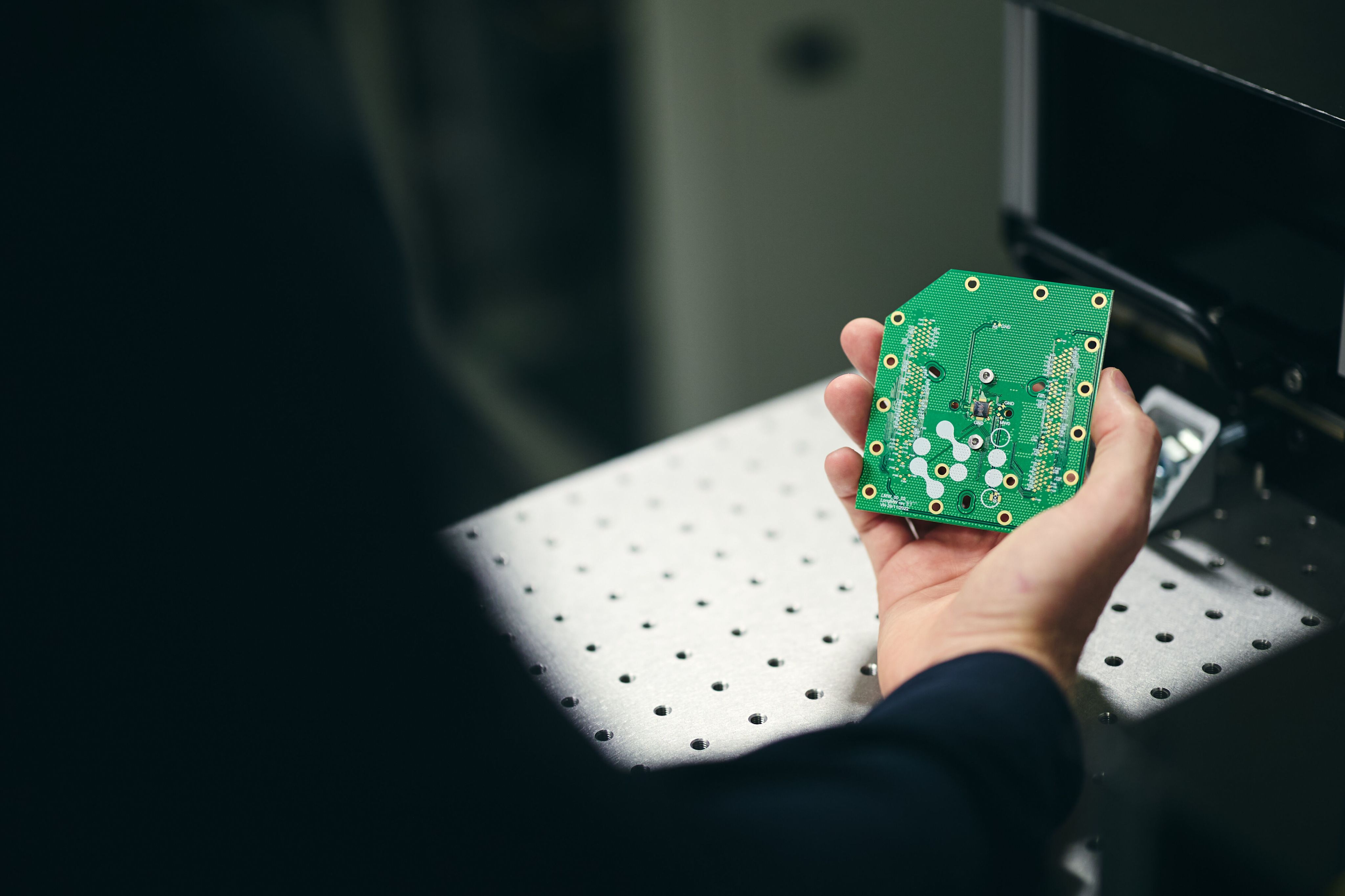 Close up of a hand holding a thin, green rectangular computer chip which has various holes and intricate patterns etched into it.