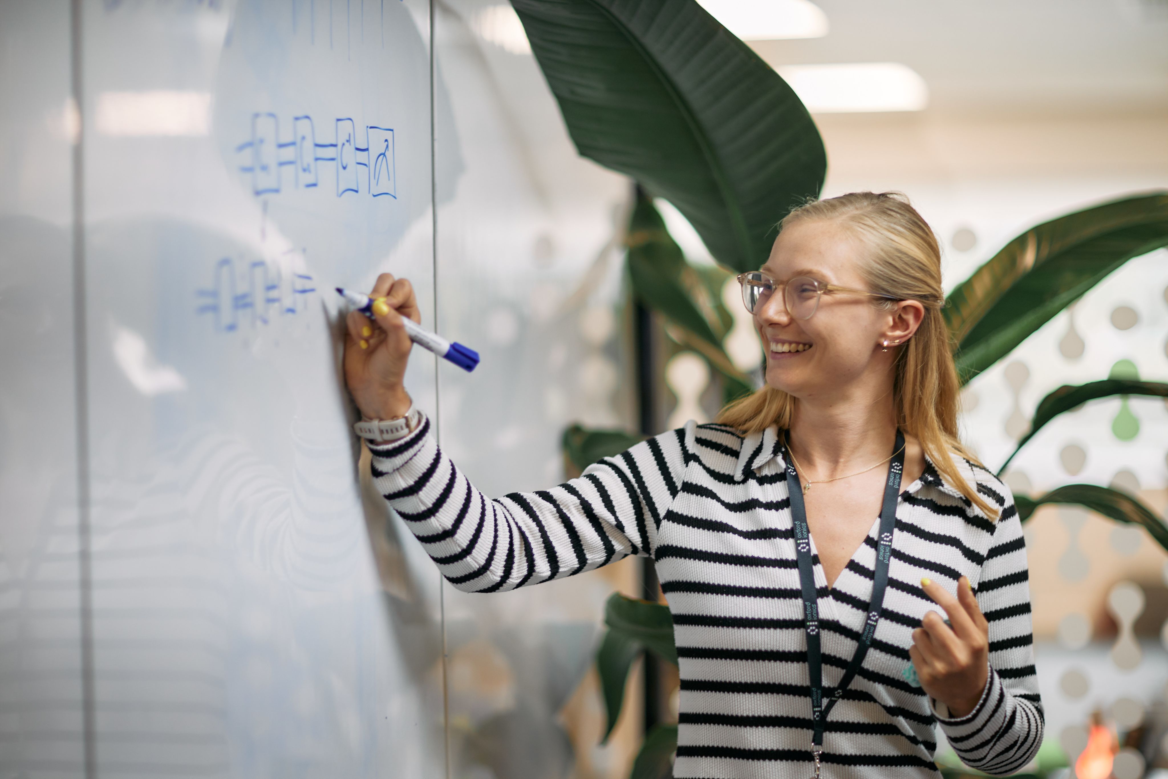 A young smiling white lady with long blonde hair and wearing glasses draws on a whiteboard in blue pen. 