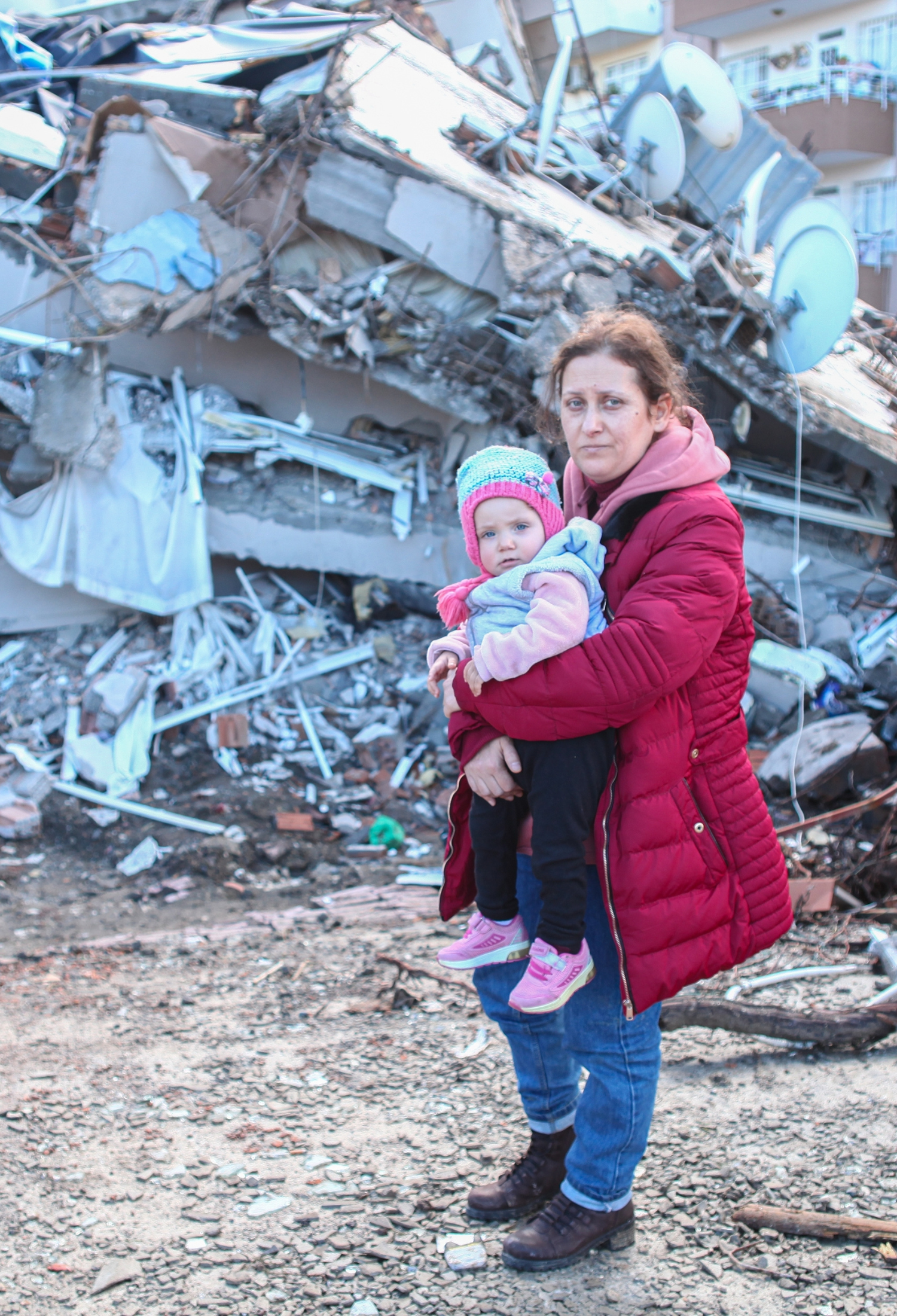 A woman holds a child in front of a collapsed building