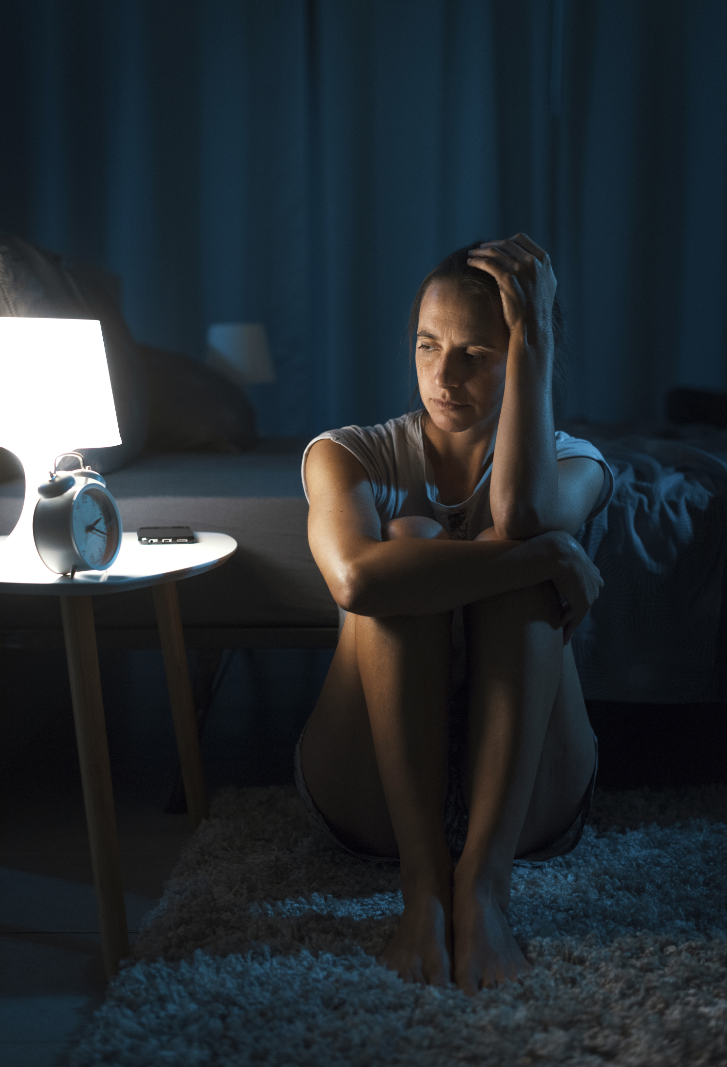 A woman sit nexts to a bed side table with the light on in the middle of the night