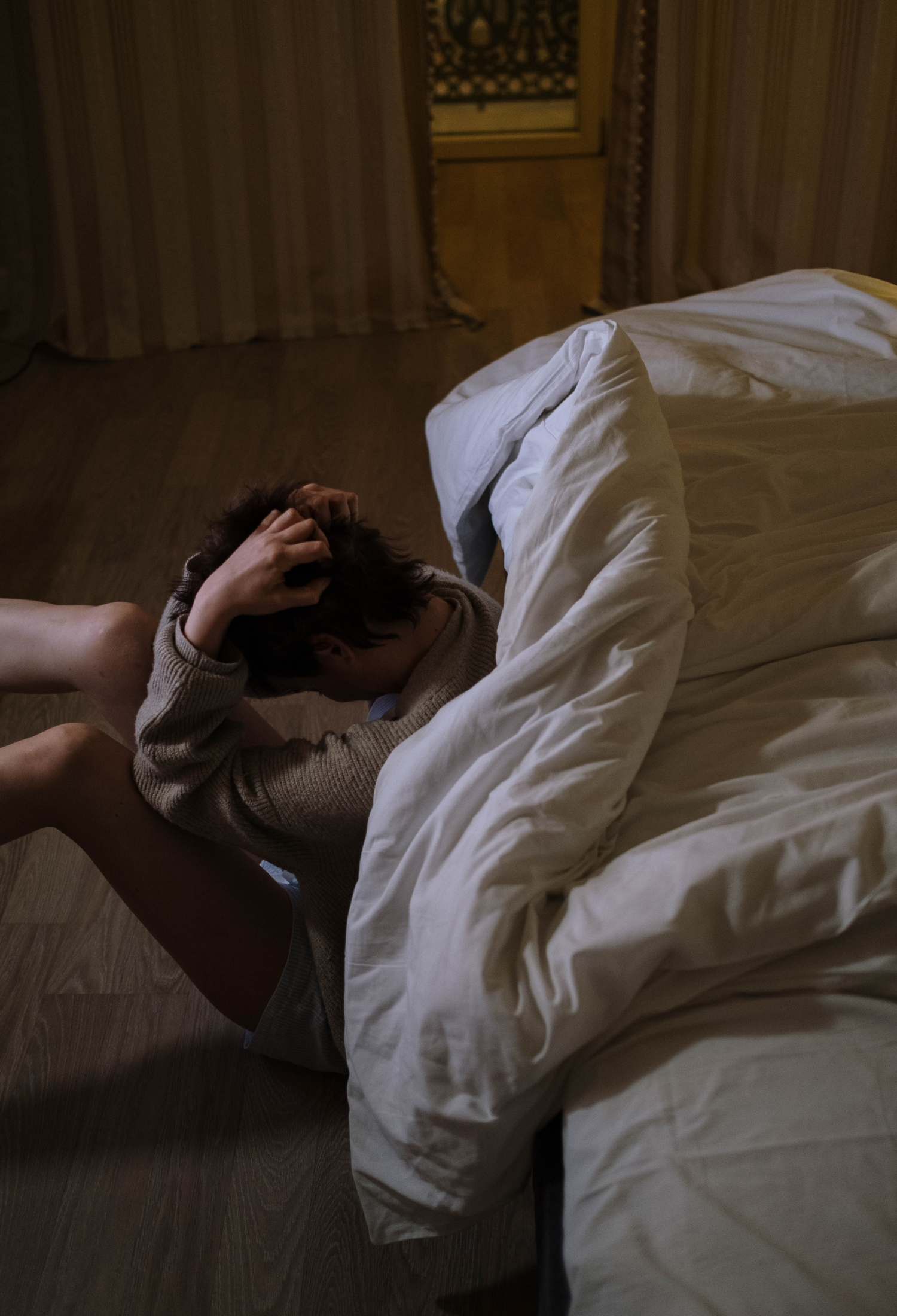 A person sits on the floor next to their bed with their head in their hands