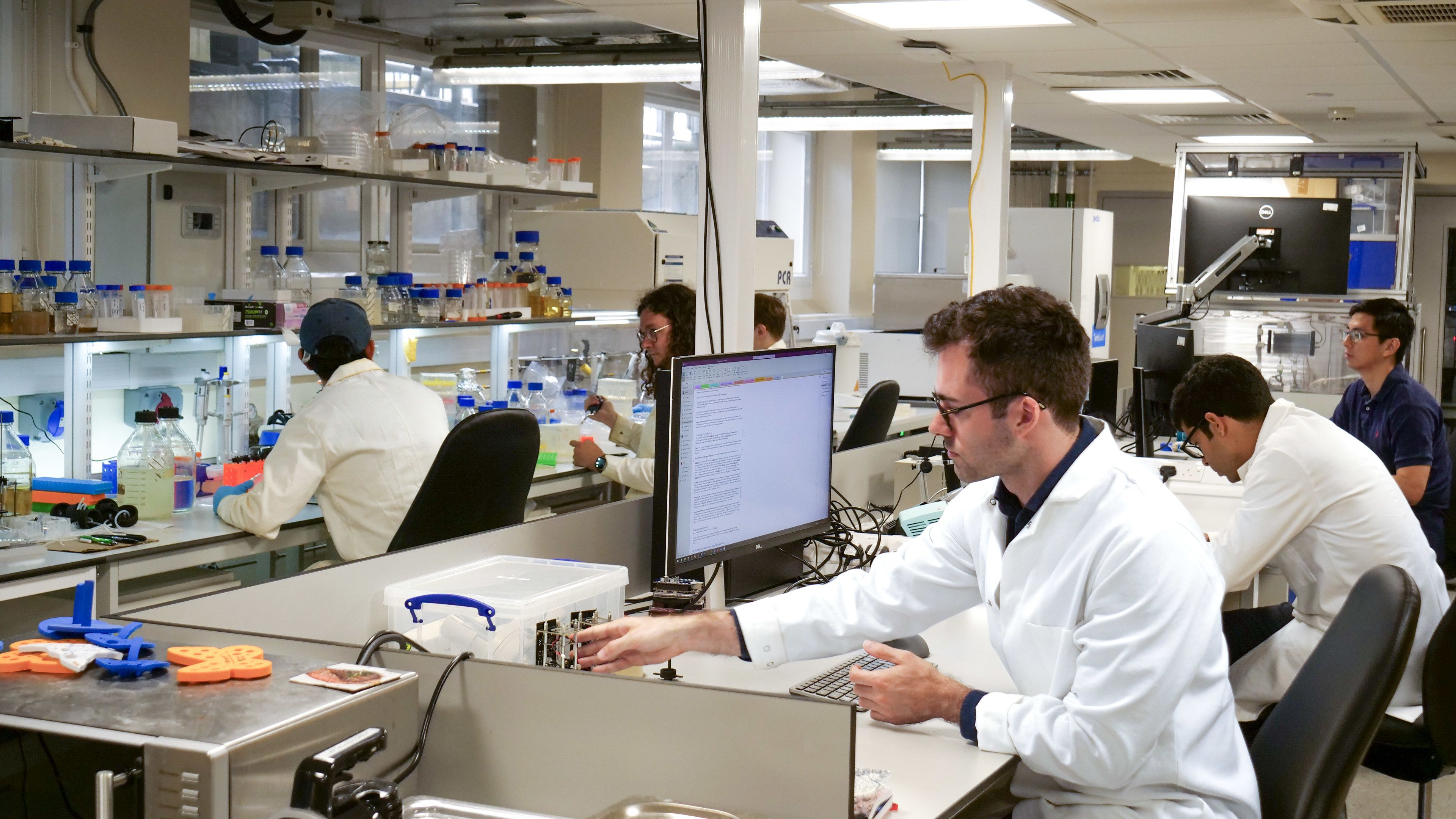 six researchers wearing lab coats sit at benches in a laboratory. Some are working at computer screens, others are carrying out experiments.