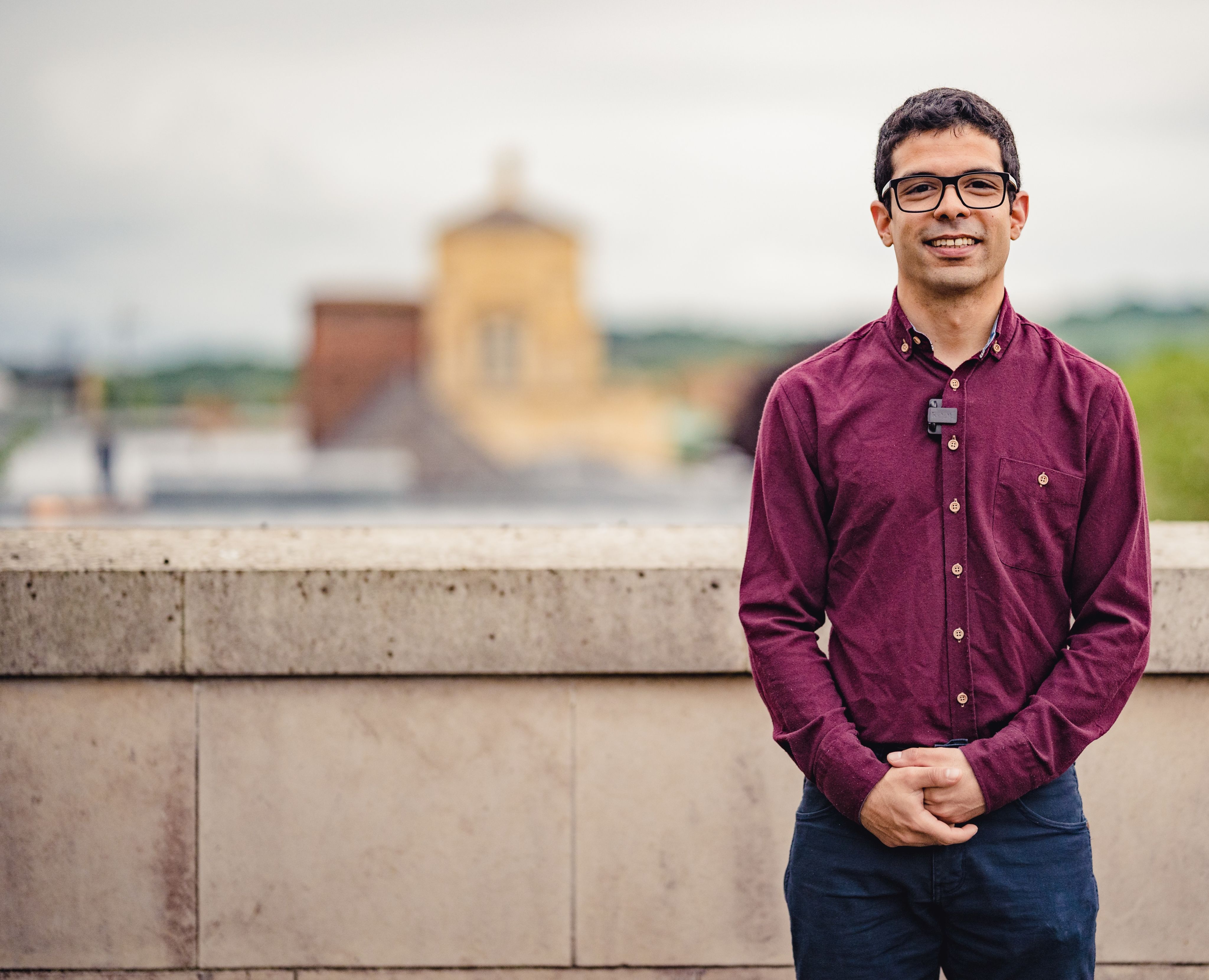 Dr Idris Kempf: a young man wearing glasses and a purple shirt. He stands in front of a stone wall with rooftops and buildings in the background.