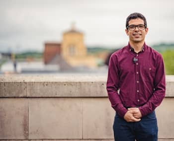 Dr Idris Kempf: a young man wearing glasses and a purple shirt. He stands in front of a stone wall with rooftops and buildings in the background.